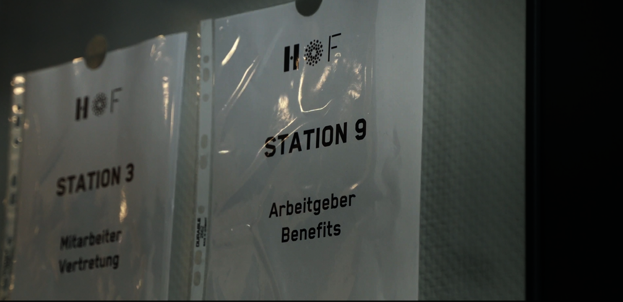 Ein DIN-A4-Blatt hängt an einer weißen Wand. Darauf ist das HOF-Logo zu sehen und der Text "Station 9: Arbeitsgeber Benefits"