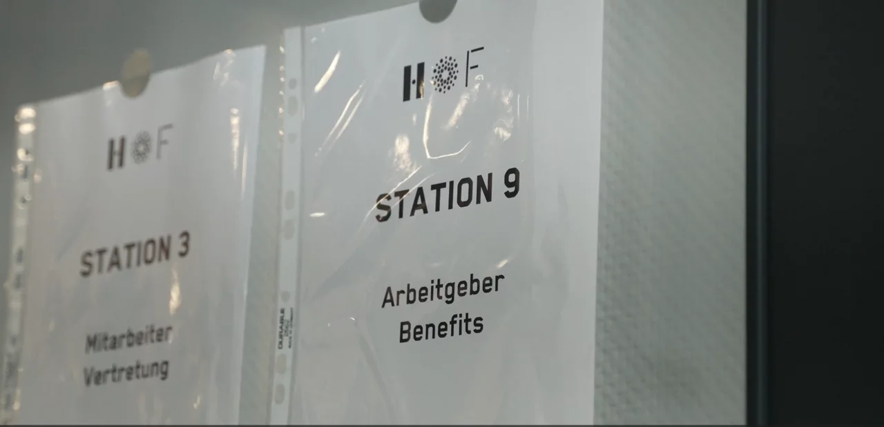 Ein DIN-A4-Blatt hängt an einer weißen Wand. Darauf ist das HOF-Logo zu sehen und der Text "Station 9: Arbeitsgeber Benefits"
