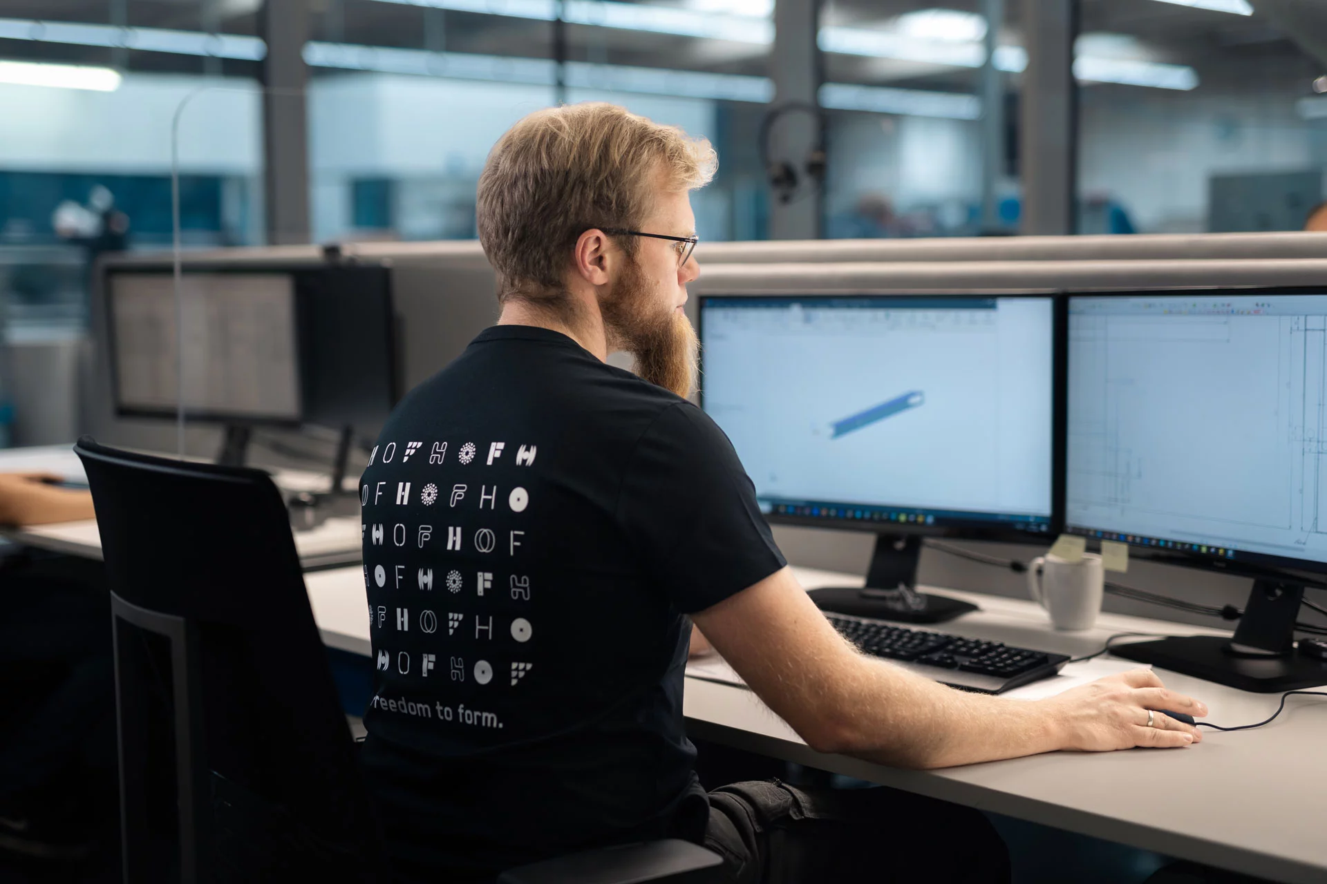 Ein junger Mitarbeiter mit roten Haaren und einem langen Bart und Brille arbeitet konzentriert an seinem Computer. Er trägt ein T-Shirt im Firmenlogo und hat ein CAD-Programm geöffnet.