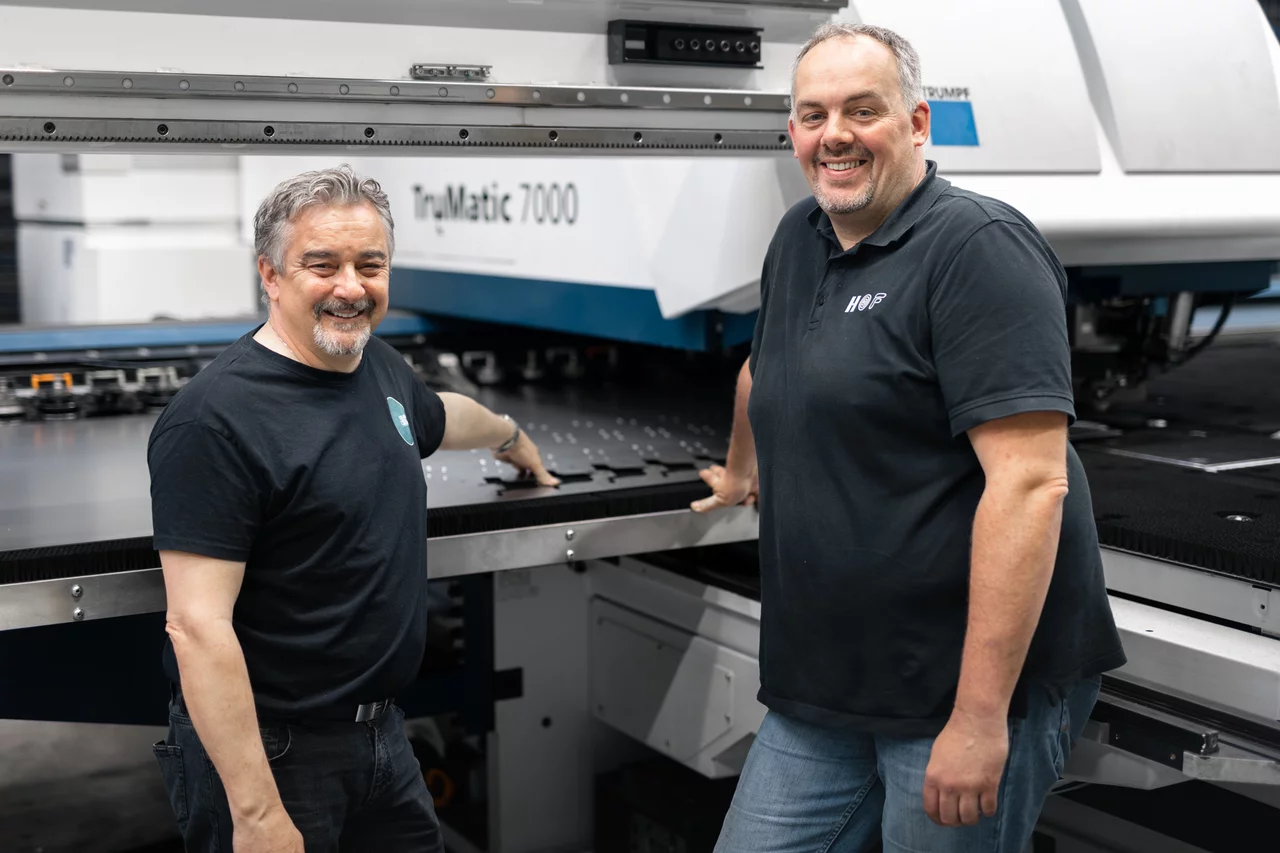 Der Gründer von GoBIC und ein Mitarbeiter von uns stehen vor einer Maschine von Trumpf und lächeln freundlich in die Kamera. Sie lehnen sich an die Maschine. Im Hintergrund ist die Bezeichnung der Maschine zu erkennen "TruMatic 7000".
