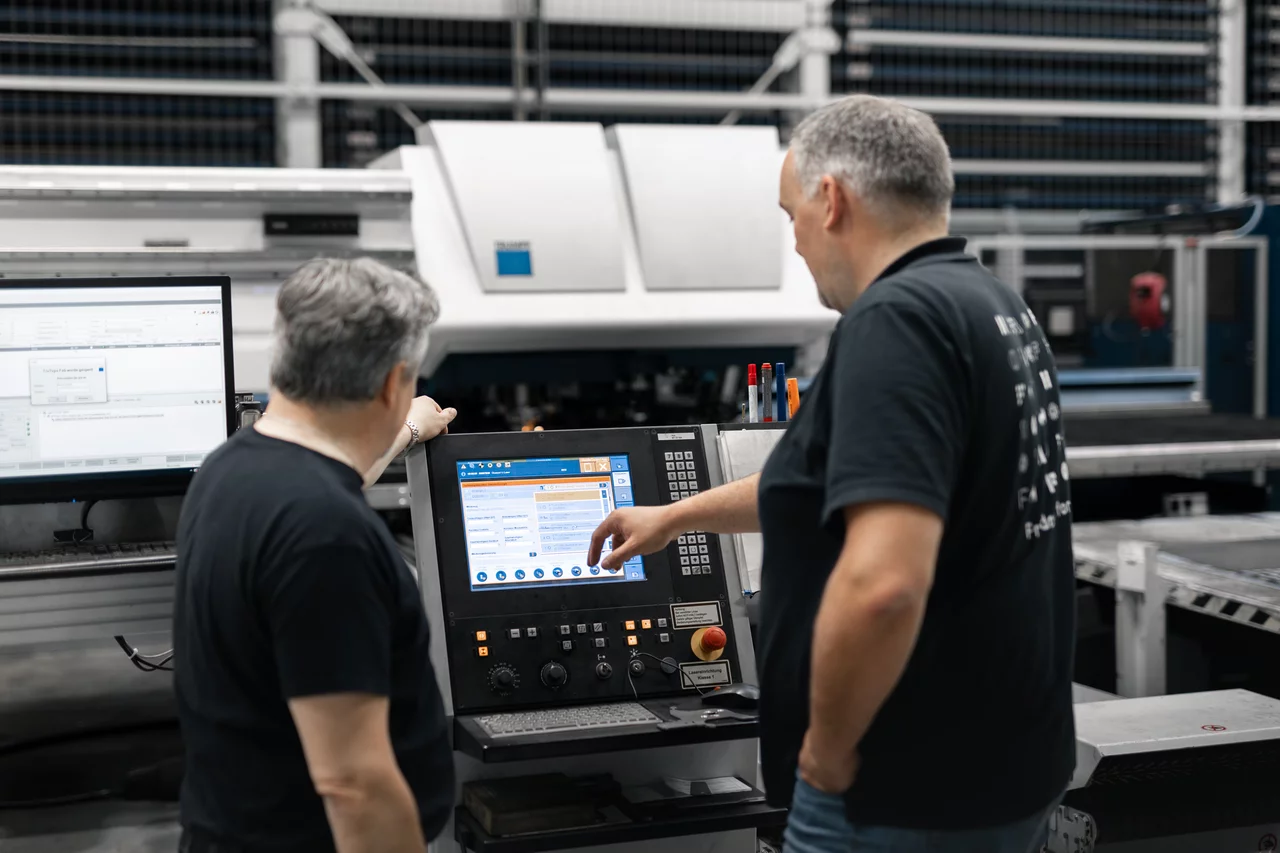 Der Gründer von GoBIC und einer unserer Mitarbeiter stehen vor dem Bedienpanel einer unserer TRUMPF-Maschinen. Im Hintergrund sieht man die Stand-Laser-Maschine von Trumpf.