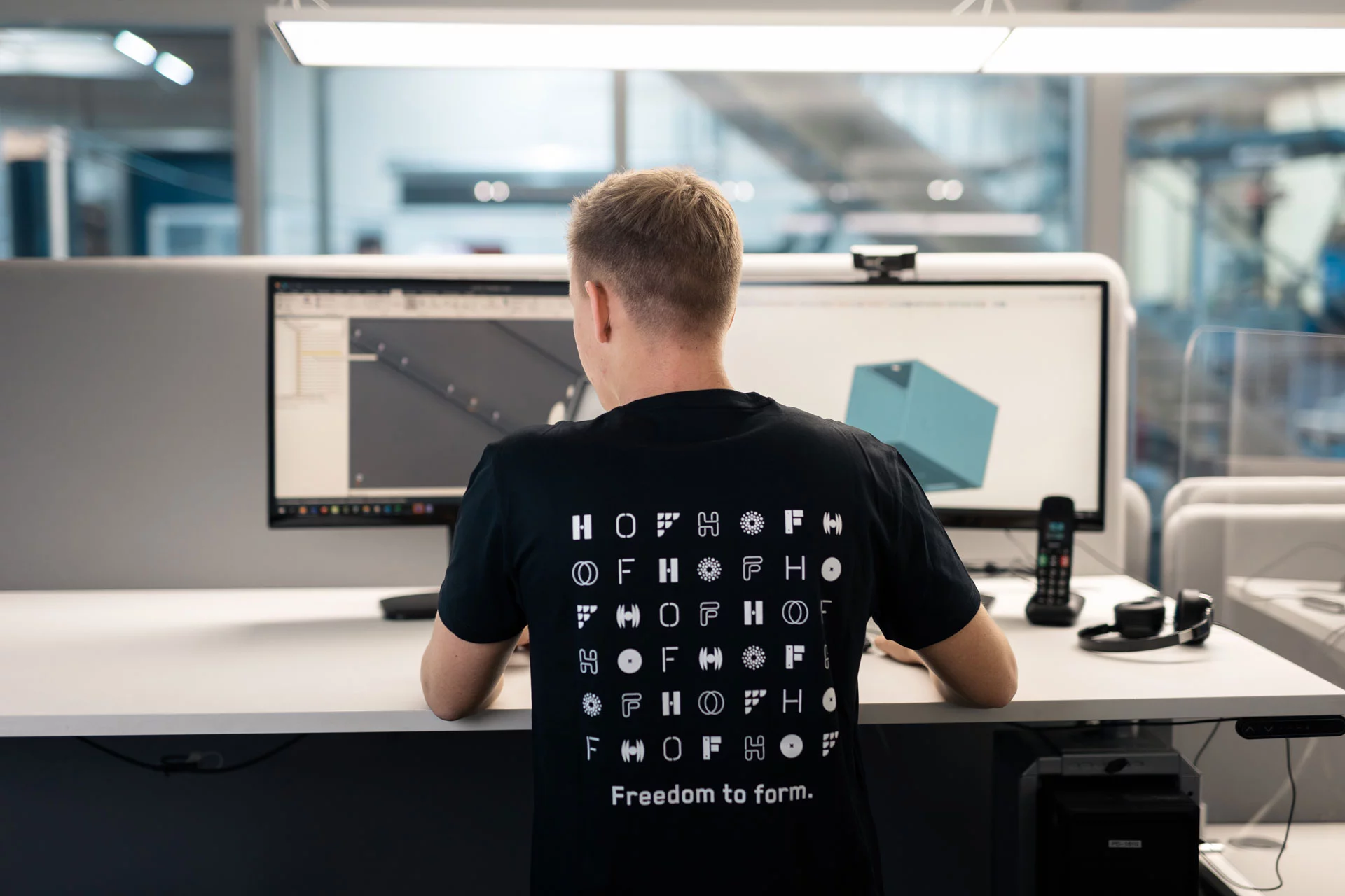 Ein junger Mitarbeiter arbeitet mit einem CAD-Programm an einem Blechteil. Er steht mit dem Rücken zur Kamera, wodurch wir einen Blick auf das Corporate Design des T-Shirts von HOF bekommen, wo ganz viele HOF-Logos zu sehen sind.