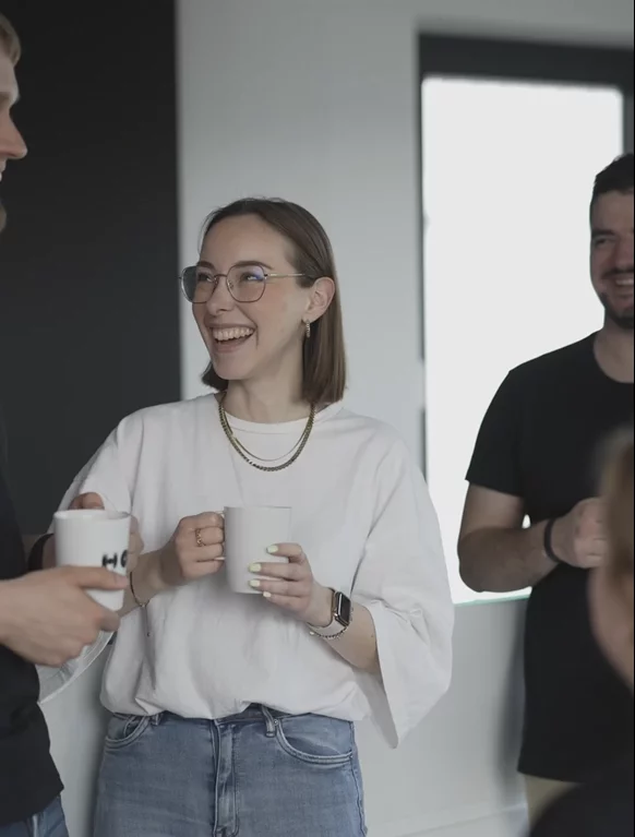 Eine junge Hof-Mitarbeiterin lacht, während sie ihren Kollegen anschaut und dabei eine Hof-Tasse in der Hand hält.