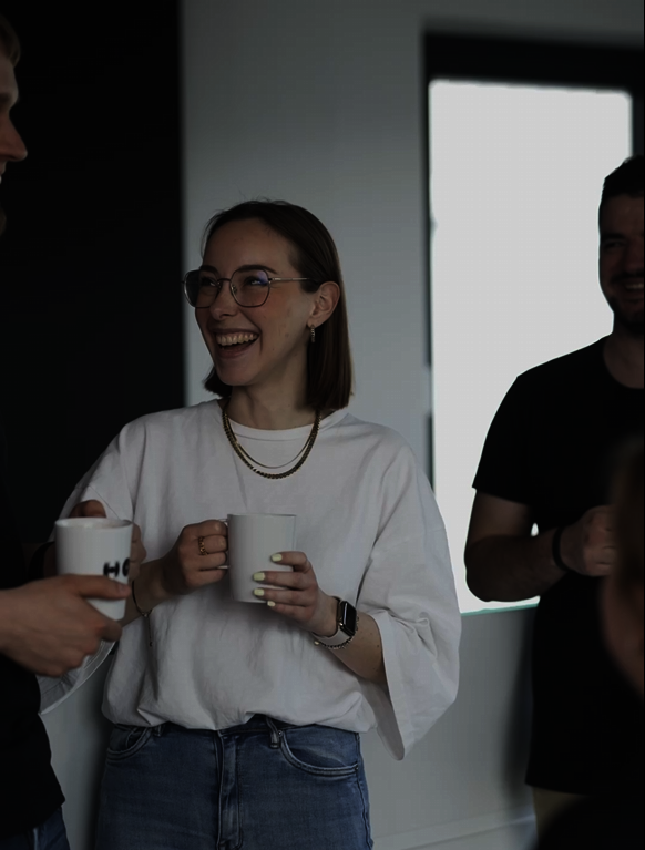 Eine junge Hof-Mitarbeiterin lacht, während sie ihren Kollegen anschaut und dabei eine Hof-Tasse in der Hand hält.