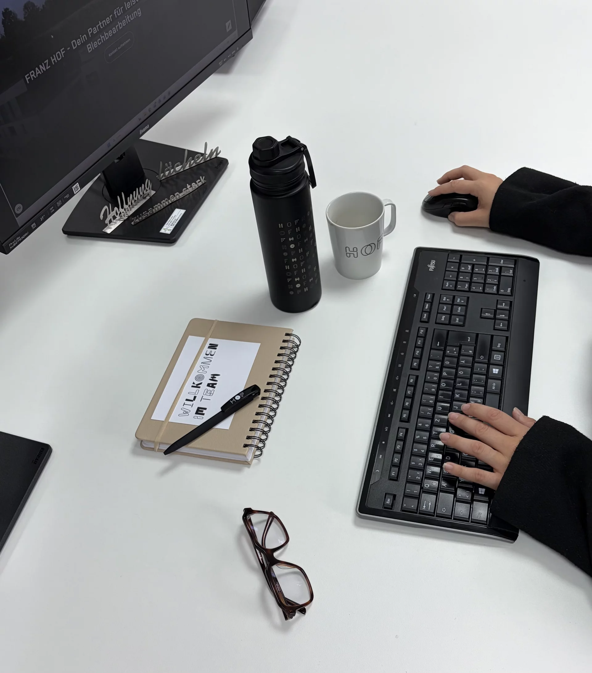 Hände an einer Tastatur an einem Arbeitsplatz mit Computer und Notizbuch