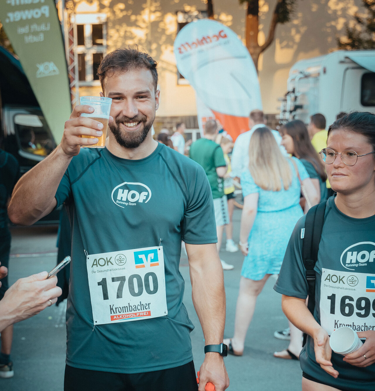 Ein HOF-Mitarbeiter prostet lächelnd in die Kamera. Er trägt seine Startnummer und ein HOF-Laufshirt.