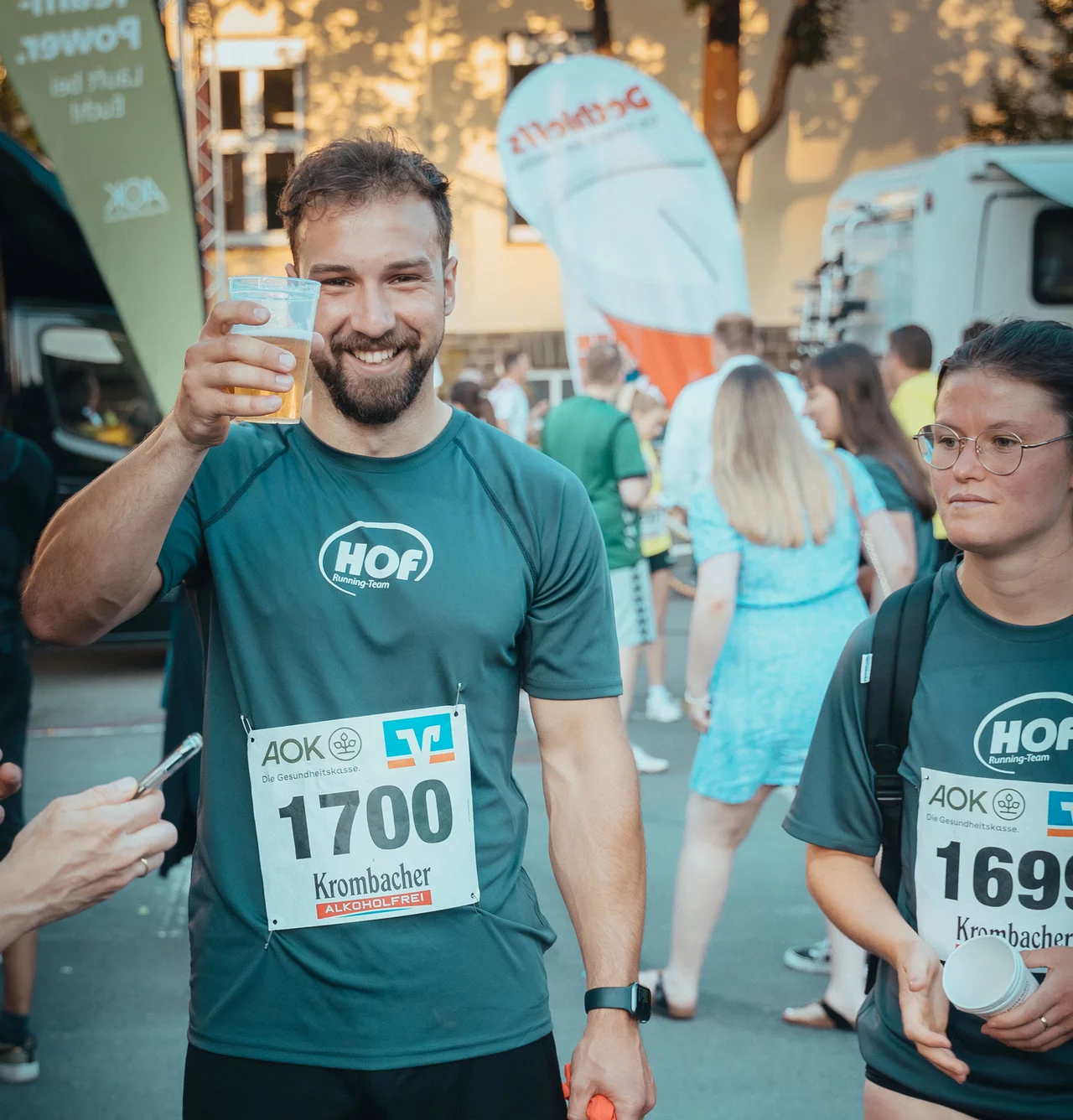 Ein HOF-Mitarbeiter prostet lächelnd in die Kamera. Er trägt seine Startnummer und ein HOF-Laufshirt.