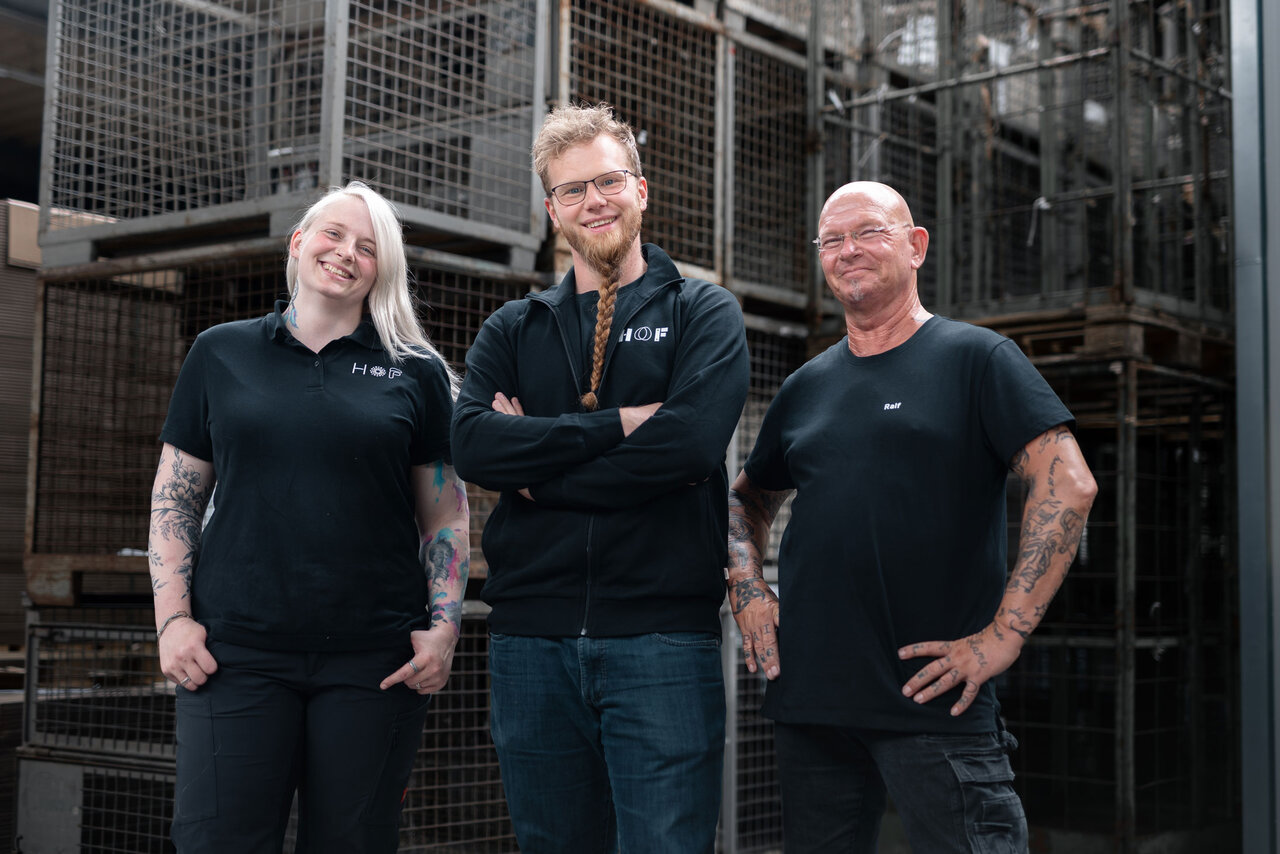 Drei Mitarbeiter präsentieren stolz ihre HOF-Kleidung. Eine tätowierte Frau (links) trägt ein T-Shirt und hat die Hände in den Hosentaschen. Ein rothaariger Mann (mittig) trägt eine Sweatjacke und hat die Arme vor dem Körper verschränkt. Ein glatzköpfiger Mann (rechts) trägt ebenfalls ein T-Shirt und hat die Hände in die Hüften gestemmt. Alle lächeln freundlich in die Kamera.