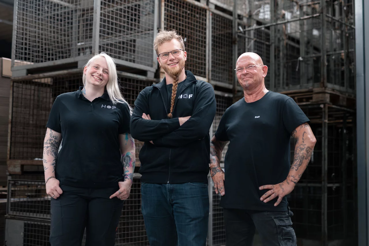 Drei Mitarbeiter präsentieren stolz ihre HOF-Kleidung. Eine tätowierte Frau (links) trägt ein T-Shirt und hat die Hände in den Hosentaschen. Ein rothaariger Mann (mittig) trägt eine Sweatjacke und hat die Arme vor dem Körper verschränkt. Ein glatzköpfiger Mann (rechts) trägt ebenfalls ein T-Shirt und hat die Hände in die Hüften gestemmt. Alle lächeln freundlich in die Kamera.