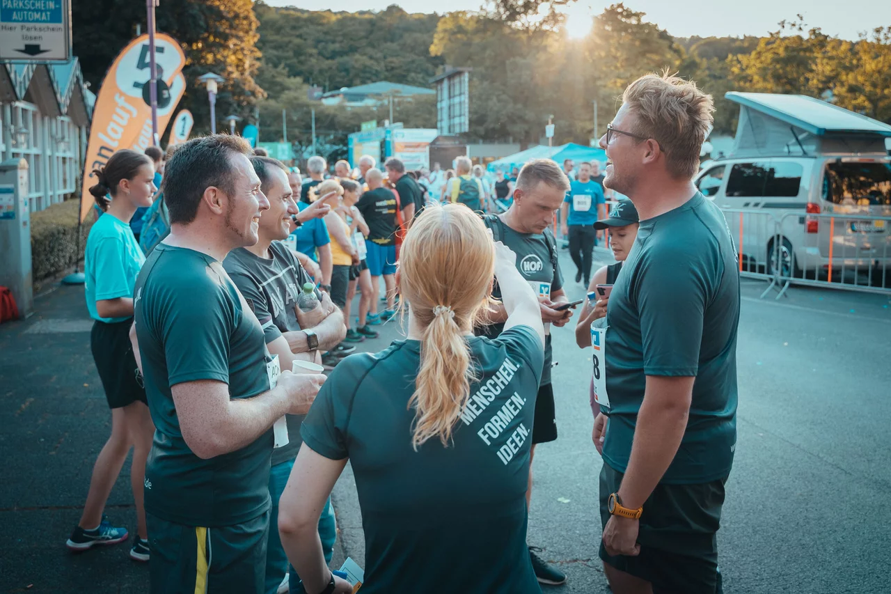 Mitarbeiter von HOF stehen in einem Kreis und unterhalten sich. Im Hintergrund sieht man das Gelände vom Firmenlauf und die tief stehende Abendsonne.