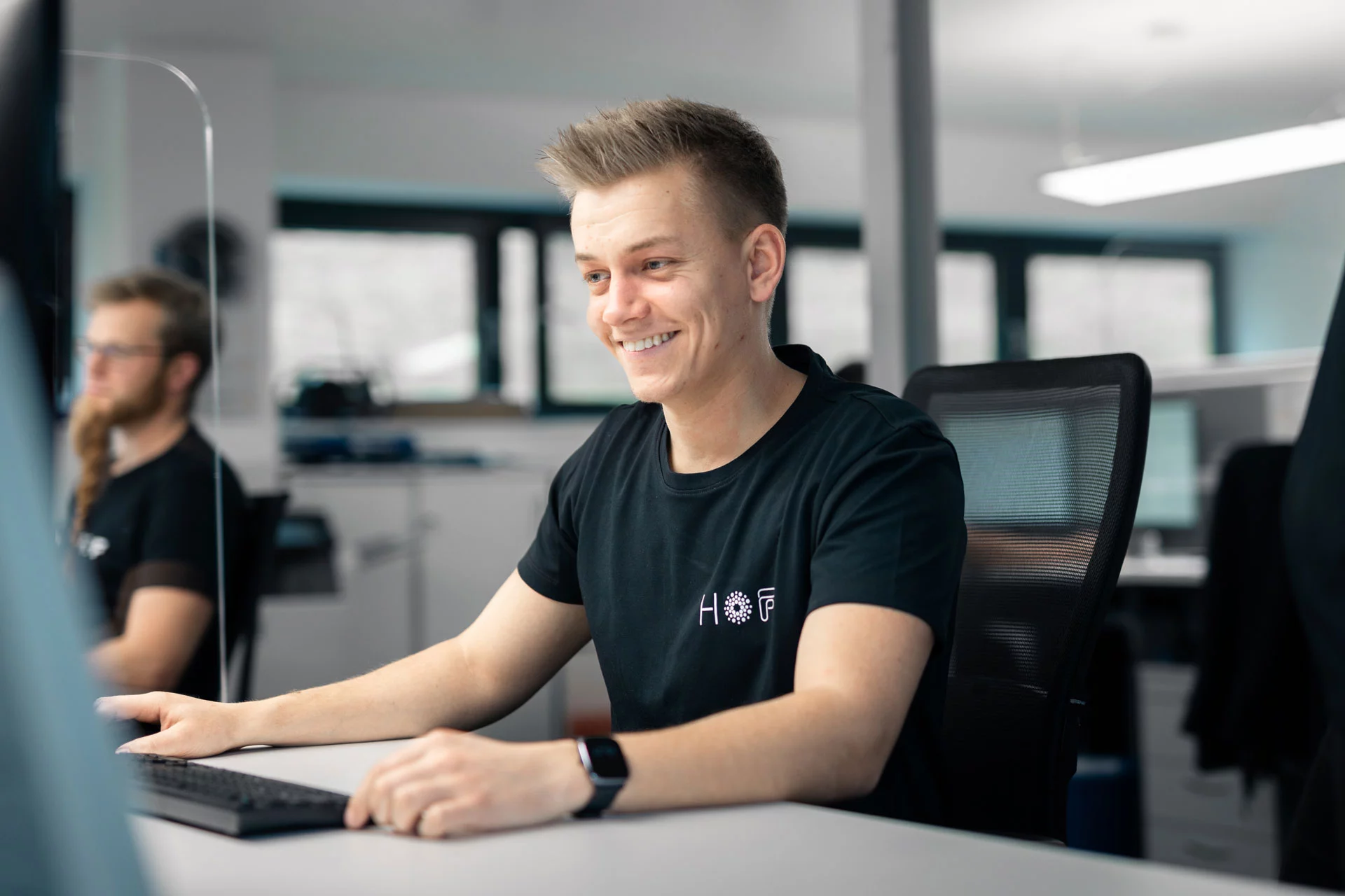 Ein junger HOF-Mitarbeiter sitzt am Schreibtisch und lächelt freundlich.