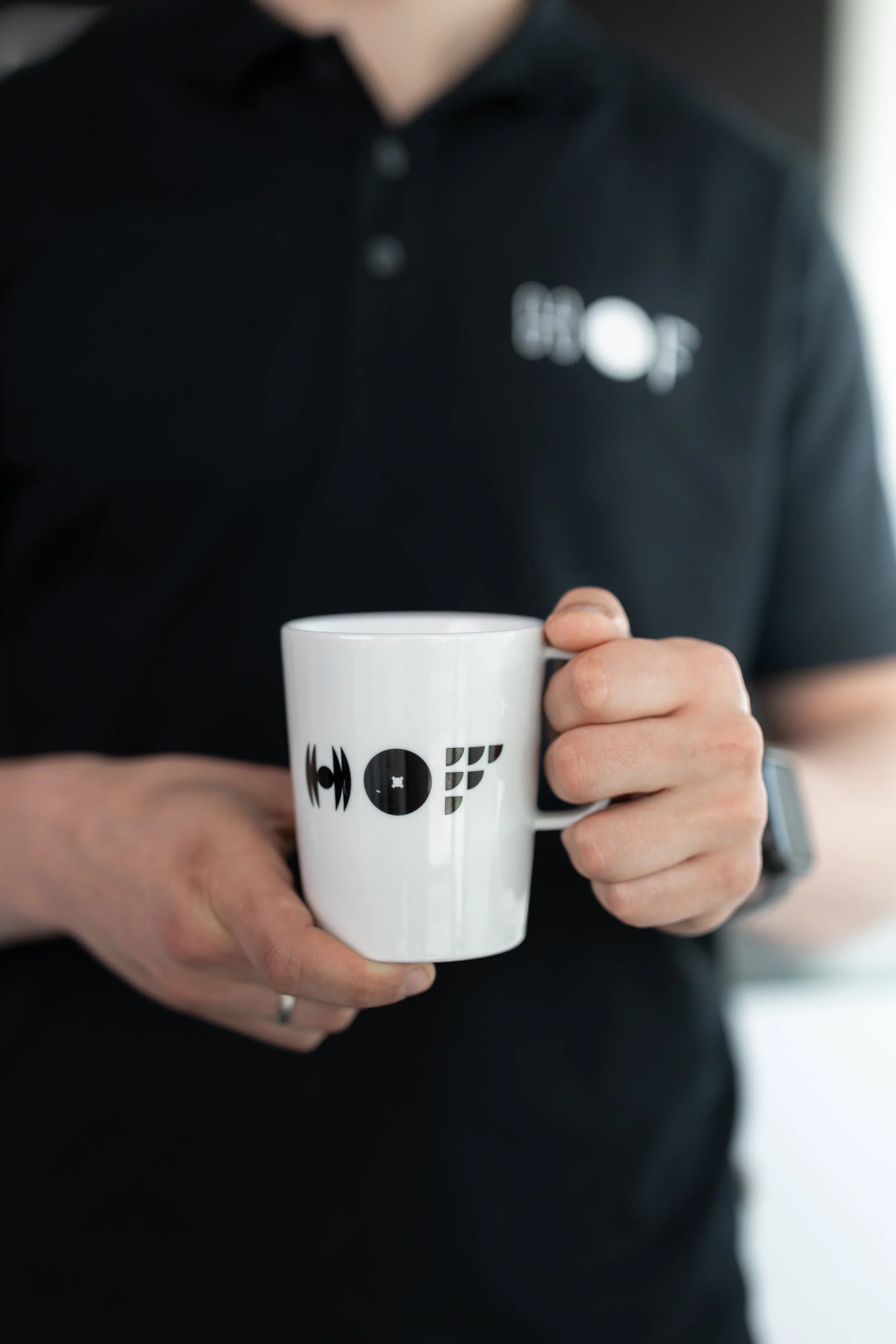 Ein Mitarbeiter von HOF mit schwarzem HOF-Shirt, hält eine weiße Tasse in die Kamera, auf der HOF steht.
