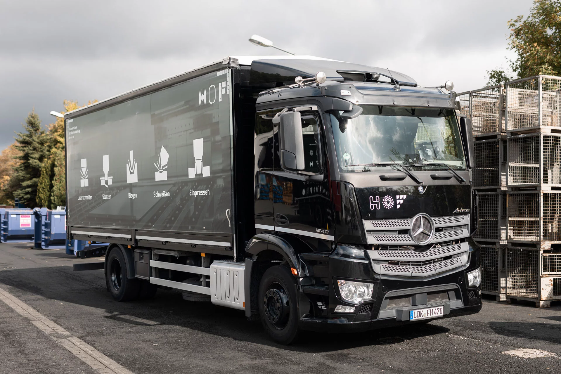 Schwarzer LKW, dessen Plane man seitlich angeschnitten sieht. Darauf zu sehen sind unsere fünf Kernkompetenzen: Lasern, Stanzen, Schweißen, Einpressen