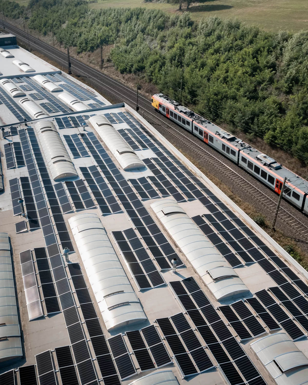 Ein Ausschnitt unserer Photovoltaikanlage. Im Hintergrund fährt vor grünen Bäumen idyllisch ein Zug vorbei.