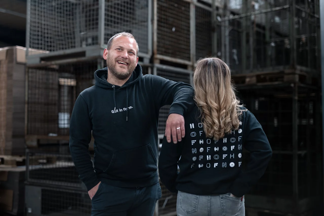 Ein HOF-Mitarbeiter (links) hat lässig seinen Arm auf den Schultern seiner Kollegin abgestützt. Seine Kollegin (rechts) steht mit dem Rücken zu uns, damit wir das Branding auf der Rückseite ihres Hoodies sehen: es sind HOF-Logos in verschiedenen Schriftarten.