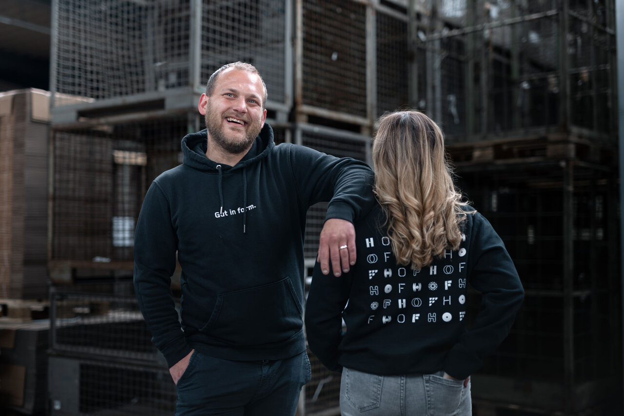 Ein HOF-Mitarbeiter (links) hat lässig seinen Arm auf den Schultern seiner Kollegin abgestützt. Seine Kollegin (rechts) steht mit dem Rücken zu uns, damit wir das Branding auf der Rückseite ihres Hoodies sehen: es sind HOF-Logos in verschiedenen Schriftarten.