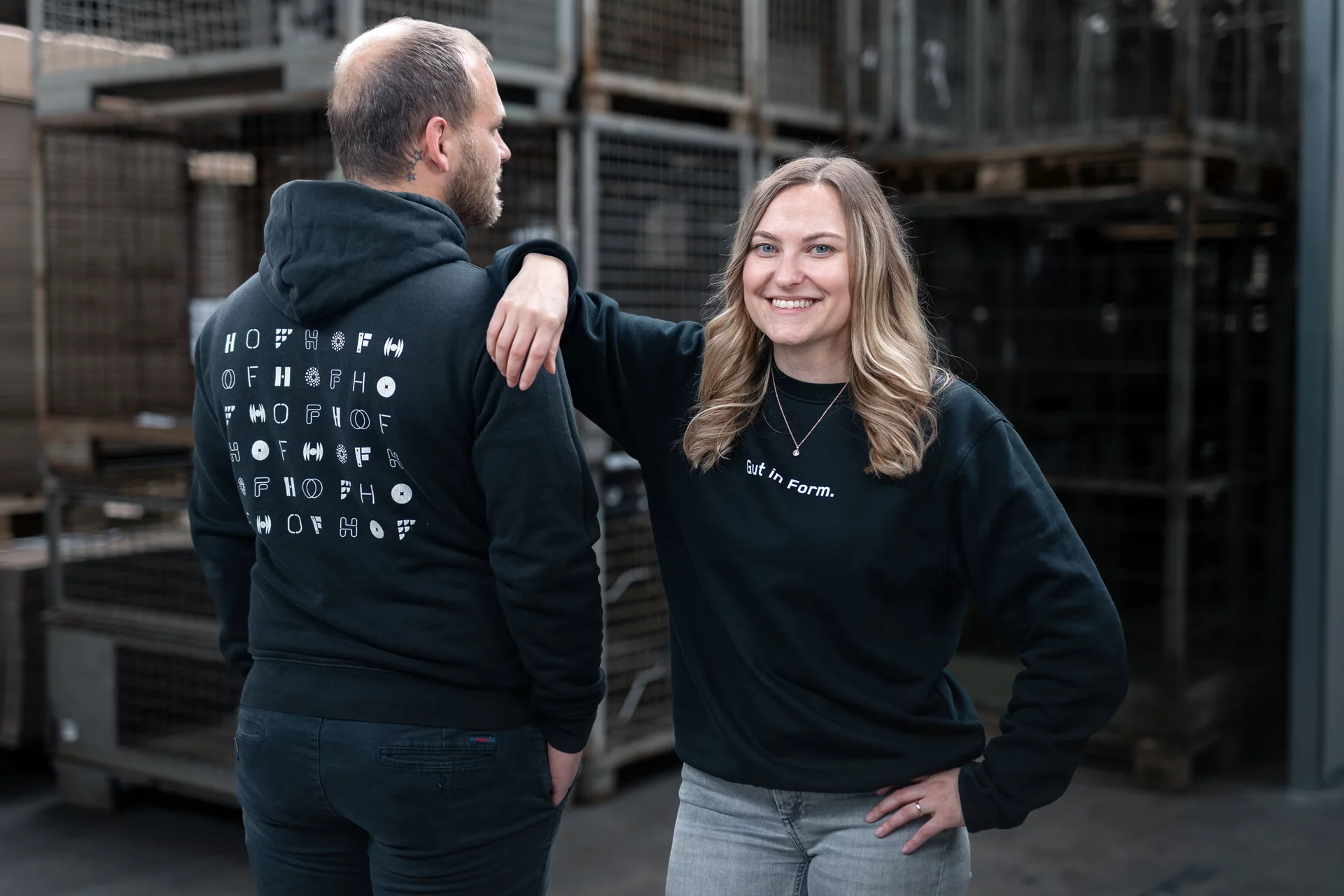 Eine HOF-Mitarbeiterin (rechts) hat lässig ihren Arm auf den Schultern ihres Kollegen (links) abgestützt. Ihr Kollege steht mit dem Rücken zu uns, damit wir das Branding auf dem Hoodie besser sehen: es sieht viele HOF-Logos in verschiedenen Schriftarten.