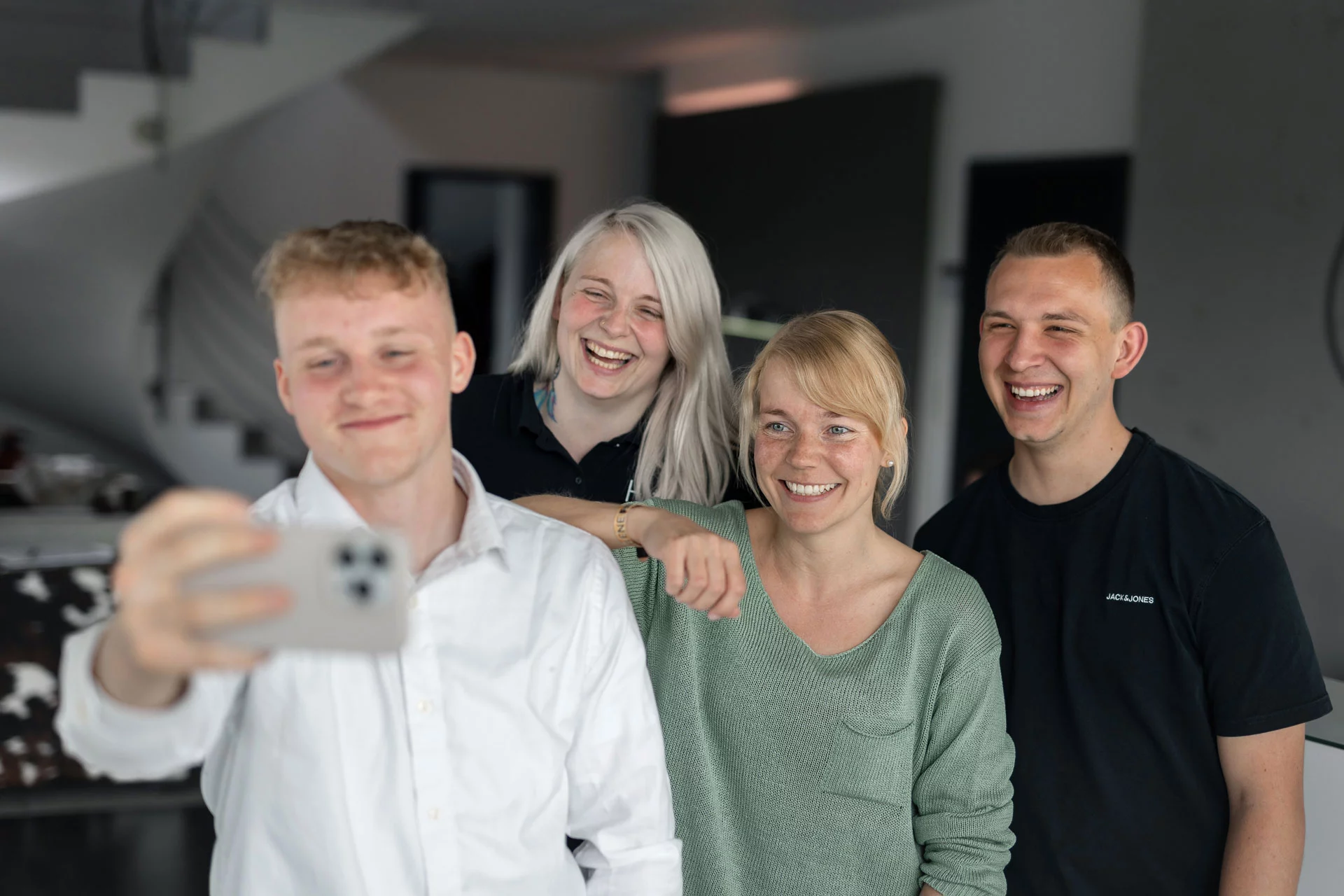 Vier junge Menschen (2 Männer und 2 Frauen) um die 20 Jahre machen ein Selfie. Der vordere Mitarbeiter hält dabei das Smartphone. Eine der jungen Frauen lehnt lässig den Arm auf seiner Schulter ab. Alle lächeln fröhlich in die Kamera. Sie sind in den Farben schwarz, weiß und mintgrün gekleidet und geben somit ein stimmiges Bild ab.