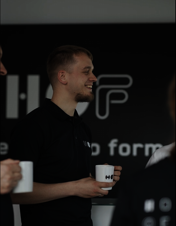 Ein junger Hof-Mitarbeiter lächelt sympathisch und hält dabei eine Hof-Tasse in der Hand.