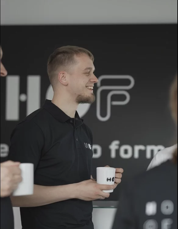 Ein junger Hof-Mitarbeiter lächelt sympathisch und hält dabei eine Hof-Tasse in der Hand.