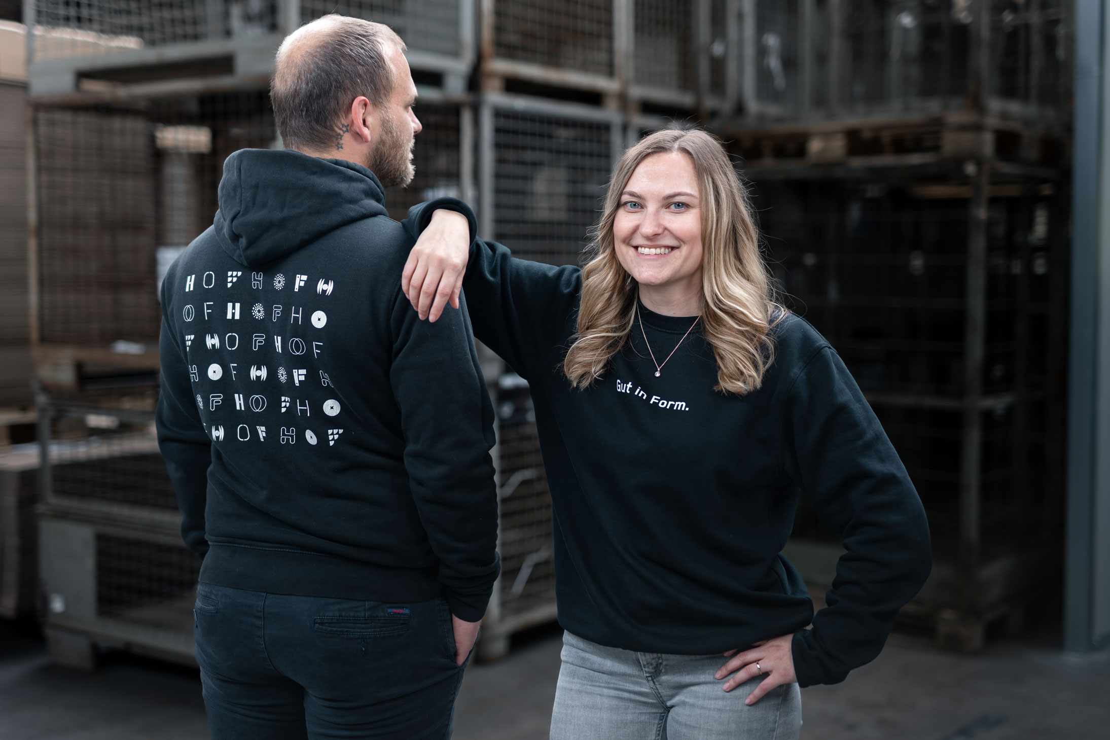 Eine HOF-Mitarbeiterin (rechts) hat lässig ihren Arm auf den Schultern ihres Kollegen (links) abgestützt. Ihr Kollege steht mit dem Rücken zu uns, damit wir das Branding auf dem Hoodie besser sehen: es sieht viele HOF-Logos in verschiedenen Schriftarten.