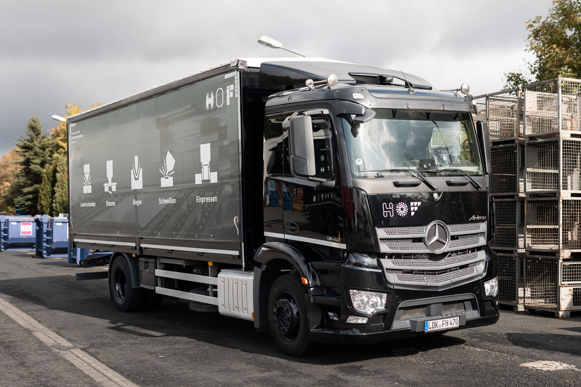 Schwarzer LKW, dessen Plane man seitlich angeschnitten sieht. Darauf zu sehen sind unsere fünf Kernkompetenzen: Lasern, Stanzen, Schweißen, Einpressen