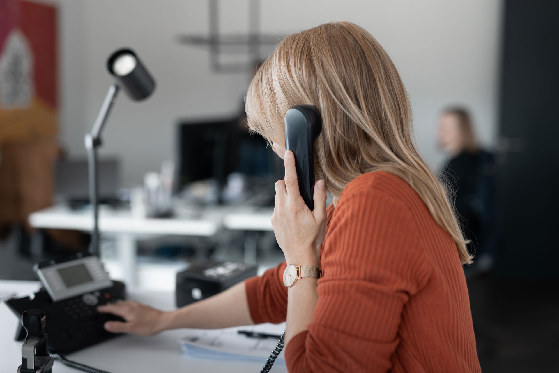 Eine blonde junge Frau hält den Hörer eines Telefons in der linken Hand und wählt mit der rechten Hand. Sie befindet sich an einem Büroarbeitsplatz.