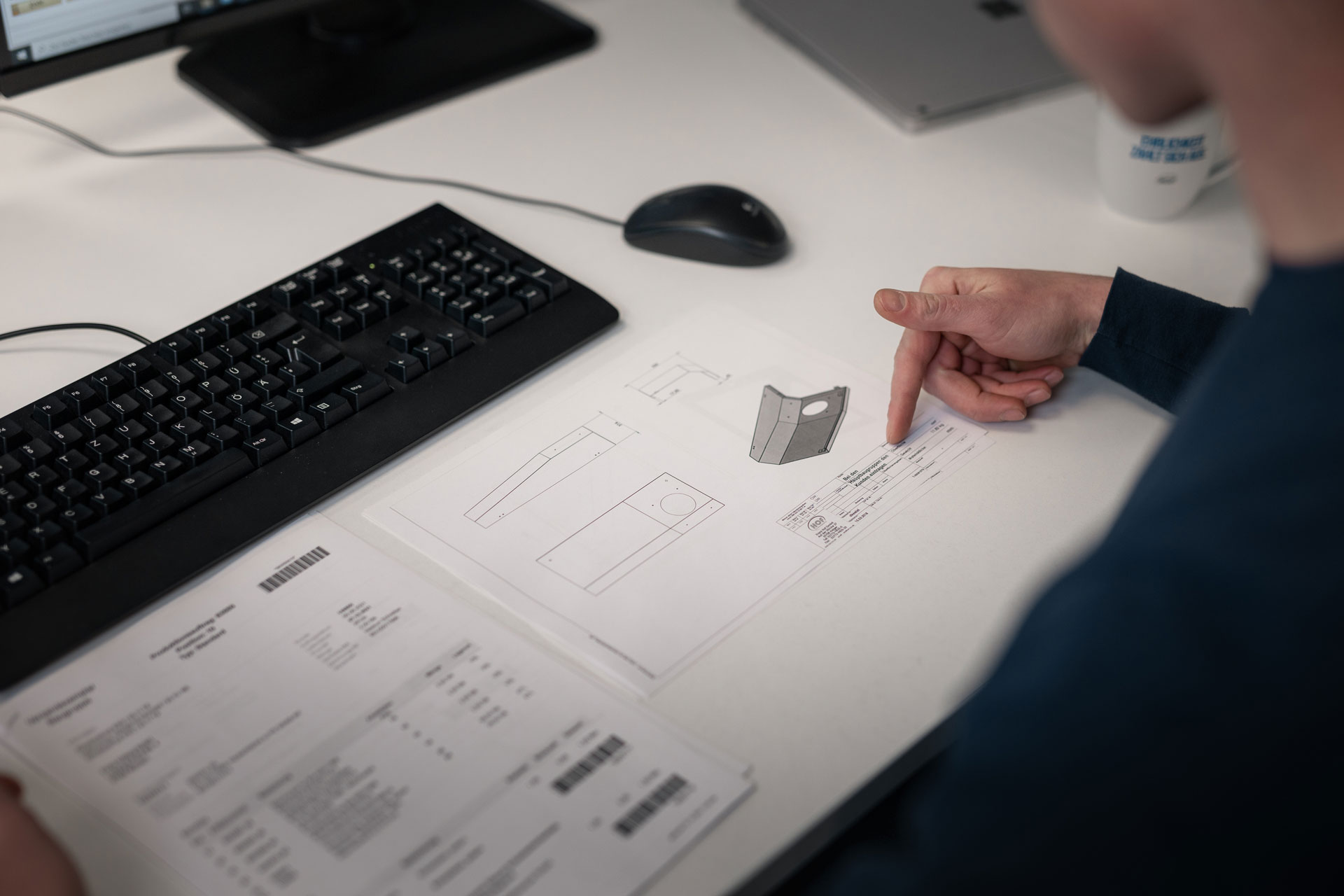 Ein Mitarbeiter sitzt an seinem Arbeitsplatz, auf dem eine technische Zeichnung liegt. Mit dem Finger zeigt er auf einen Teil der technischen Zeichnung.
