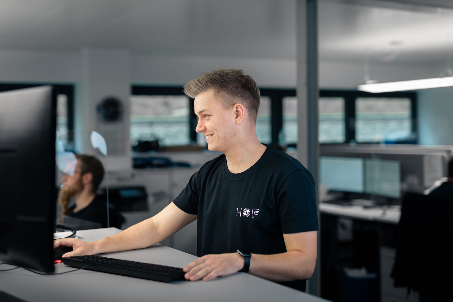 Ein junger Mitarbeiter sitzt vor seinem PC, mit einer Hand an der Maus und der anderen auf der Tastatur. Er schaut auf den Monitor, lächelt zufrieden und trägt ein T-Shirt von HOF.